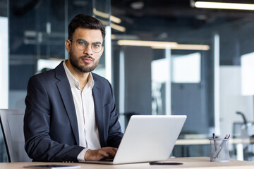 A confident businessman in a navy suit and glasses, working on a laptop in a modern office setting.