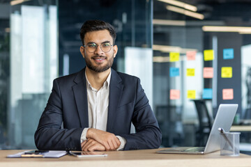 A smiling businessman in a modern office setting, seated at a desk, looking directly at the camera.