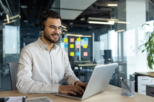A smiling man with glasses and a headset works on a laptop at an office desk. He appears to be engaged in a video call. - Powered by Adobe