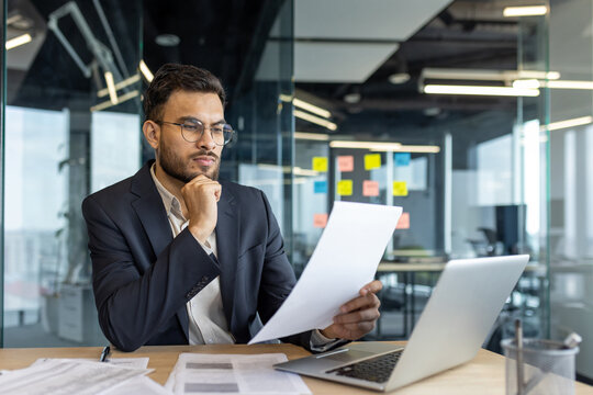 A thoughtful businessman examines documents in his office. A laptop sits on the desk. - Powered by Adobe