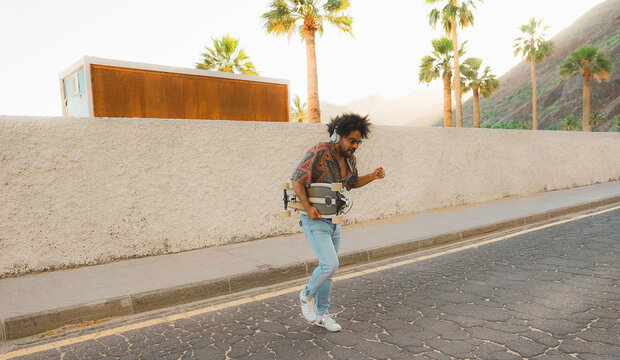 African man dancing with palms and beach in background - Afro skater having fun outdoor listening music with headphone set - Vintage filter and summer lifestyle concept - Main focus on face - Powered by Adobe