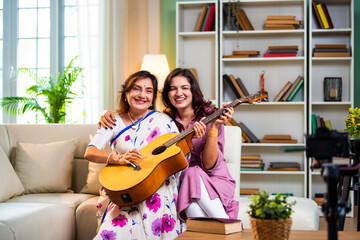 Indian daughter teaching guitar to elderly mother while seated together at home indoors
