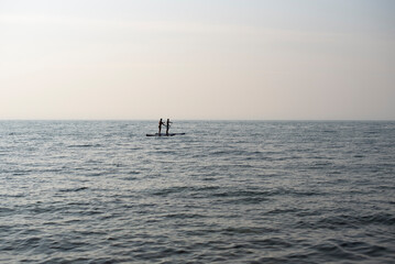 Fototapeta premium Two people stand on a paddleboard in calm ocean waters. The scene is serene with a clear sky and gentle waves.