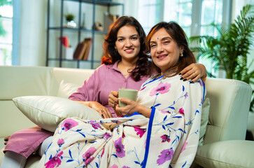 Indian mother and daughter relaxing on sofa and talking over hot coffee in cozy modern home