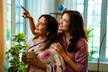 Indian mother daughter enjoy morning tea by window while smiling and bonding peacefully at home