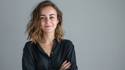 Confident young woman with short brown hair wearing black shirt standing with arms crossed and smiling at the camera on a grey background