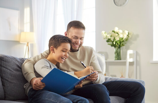 Caring young father helps his little son with doing homework task, writing something down in notebook, sitting together on comfortable sofa and embracing. Fatherhood and family concept - Powered by Adobe
