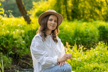 A young woman in a light shirt and hat sitting on a tree stump by the river surrounded by greenery. She holds glasses in her hands, which adds naturalness and harmony to the composition.