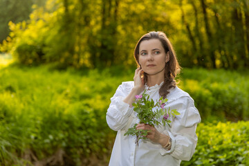 a woman in a white shirt with ruffles, holding a bouquet of green leaves and flowers. It stands...