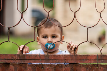 Toddler with pacifier looking through rusty metal fence