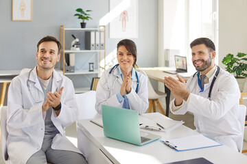 Fototapeta premium Happy smiling doctors applauding on a meeting sitting at the desk on workplace in clinic. Successful team of young medical staff sitting in hospital on conference. Medicine and health care concept.