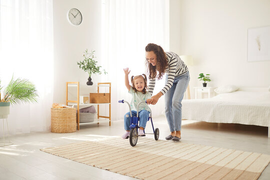Portrait of a cute little child girl riding a bike with mother in living room at home together. Happy young woman parent playing with daughter teaching her to ride a bicycle. Family leisure concept.