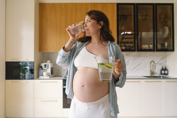 In a contemporary kitchen, a pregnant woman with a joyful expression holds a pitcher filled with water and a glass. Bright light enhances her radiant smile as she enjoys hydration.
