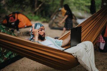 A man lying in a hammock in a forest campsite, using a laptop. There are tents and people in the...