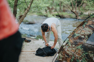 A man prepares or organizes his backpack while setting up a camping site near a forested river. The...
