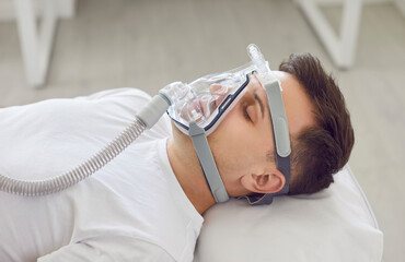 Unconscious male patient lying on bed with oxygen mask on his face during treatment in hospital ward. Sick young man wearing aspirator sleeping on bed in intensive care unit in medical clinic.