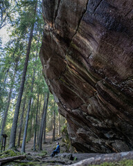 Dramatic rock formation at Paistjärvi’s Pirunkirkko (Devil’s Church), a natural cliff in a Finnish forest. A person sits below the massive boulder, highlighting the scale of the geological feature. 