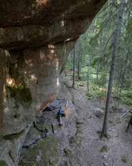 View from above of a man sitting on a bench under the towering rock formation at Pirunkirkko (Devil’s Church) in Paistjärvi, Finland. The scene captures the scale of the granite cliff.