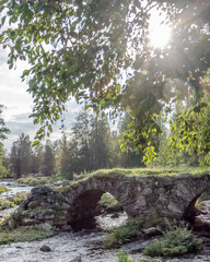 A rustic stone arch bridge spans a gently flowing river in the Finnish countryside, with sunlight filtering through green tree leaves. The peaceful rural scene evokes timeless natural beauty.