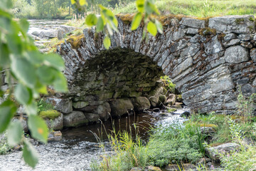 A detailed view of a historic stone arch bridge with moss-covered rocks and flowing water beneath. The structure showcases traditional craftsmanship and blends naturally into the lush greenery.