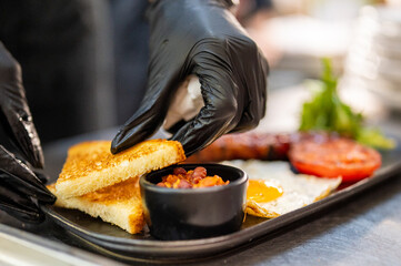 Chef in black gloves plating gourmet breakfast with toast, beans, fried egg, grilled tomatoes, and...