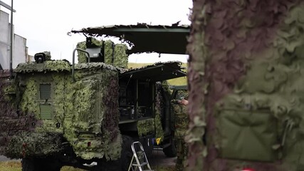 Camouflaged military communication vehicle with open panels and exposed control systems ready for field deployment as soldier adjusts setup in tactical command zone