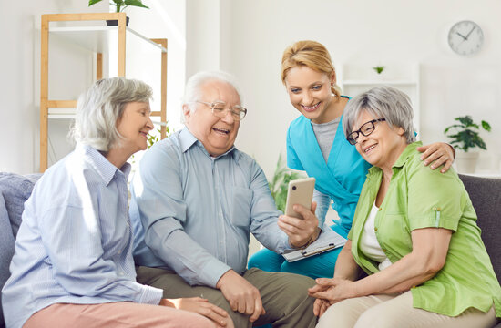 A group of happy elderly people making a video call from a smartphone in a guest house. Nurse and senior people laugh while looking at the smartphone screen. Concept social connection