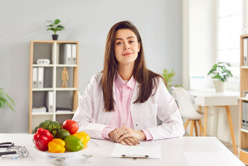 Portrait of cheerful young friendly female doctor nutritionist in medical uniform sitting on...