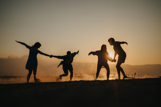A group of friends plays and dances on the beach during a captivating sunset, silhouetted against the golden light, evoking feelings of joy, unity, and the beauty of shared moments.