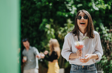 A cheerful woman, dressed casually, smiles under the sunlight while enjoying a refreshing drink...