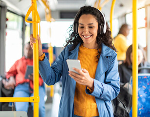 A woman is on a bus with a cell phone in her hand. She is smiling and wearing headphones