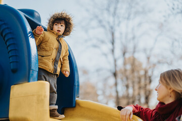 A young child wearing a warm jacket with a hood stands on a colorful playground structure, looking...