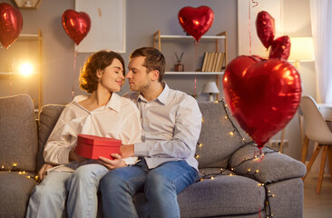 Happy loving couple hugging, kissing, and holding a gift during a Valentine Day date at home. Love,...