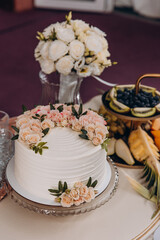 Single-tier white wedding cake decorated with a semicircle of peach and blush roses and green leaves. Displayed on a vintage white table alongside champagne, fruit platter, floral bouquet, and elegant