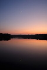 Obraz premium Serene Sunset over Calm Lake with Crescent Moon Reflection