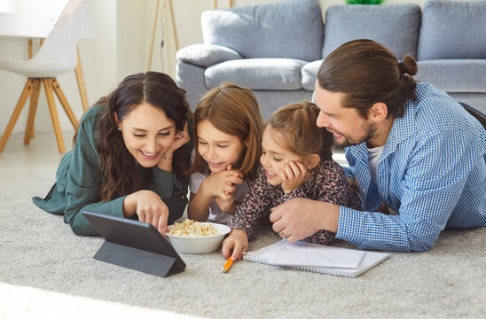 Happy young family lying on carpet, watching something funny on tablet screen together while eating popcorn at home. Relaxed cheerful parents and kids enjoying online leisure time in living room. - Powered by Adobe