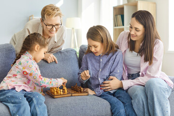 Positive kids with grandmother and mother enjoy leisure time playing chess at home, focusing on...
