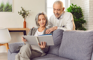 Elderly couple is happily looking at a family photo album together, sitting on sofa at home. Joyful...