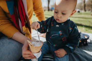 A child sits on a picnic blanket outdoors, tasting ice cream from a cone with their family present....
