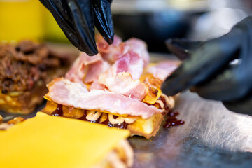 Close-up of gloved hands preparing a waffle sandwich with bacon, melted cheese, and sauce. Street food preparation in progress, emphasizing texture, freshness, and appetizing detail.