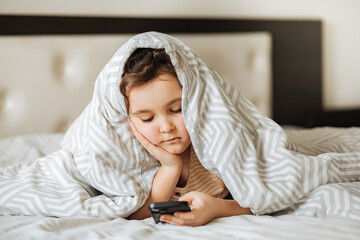 Cute little kid girl using smart phone lies on bed under the cover at home