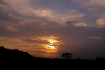 A beautiful sunset with vibrant orange hues, silhouetted by treetops and hills, creates a serene evening scene.