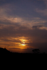 A beautiful sunset with vibrant orange hues, silhouetted by treetops and hills, creates a serene evening scene.