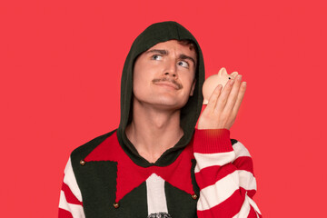 Man wearing a festive sweater with a hood holds a small piggy bank while looking thoughtfully...
