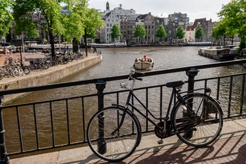 Fototapete A black bicycle parked on a bridge, offering a view of an Amsterdam canal with a small boat and distant buildings © Yevhenii Rukavitsyn