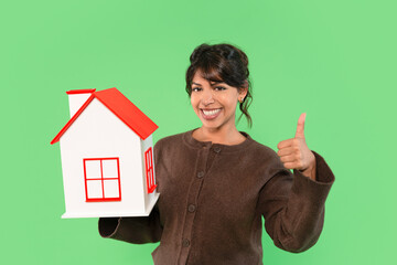 Woman smiles and gives thumbs up while holding a small model house in front of a green background