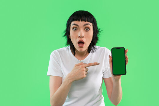 Excited woman points at smartphone screen while standing in front of a bright green background during a casual moment