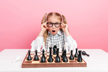 Smart young schoolgirl in glasses and formal dress surprised while playing chess on a pink background