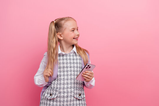 Cheerful schoolgirl in stylish school uniform holding smartphone, ponytail hairstyle, pink background, formalwear concept - Powered by Adobe