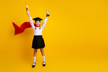 Cheerful schoolgirl in academic attire holding diploma and trophy against vibrant yellow backdrop...